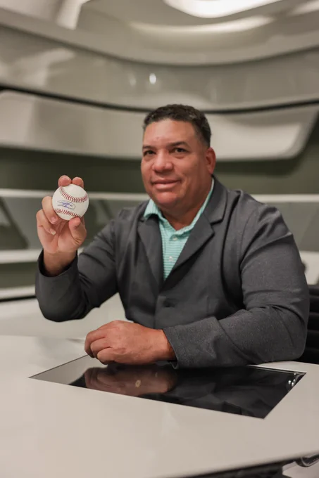 Bartolo Colon holds an autographed baseball in his right hand as he smiles for a posed photo. He wears a gray buttoned jacket over a green striped polo.