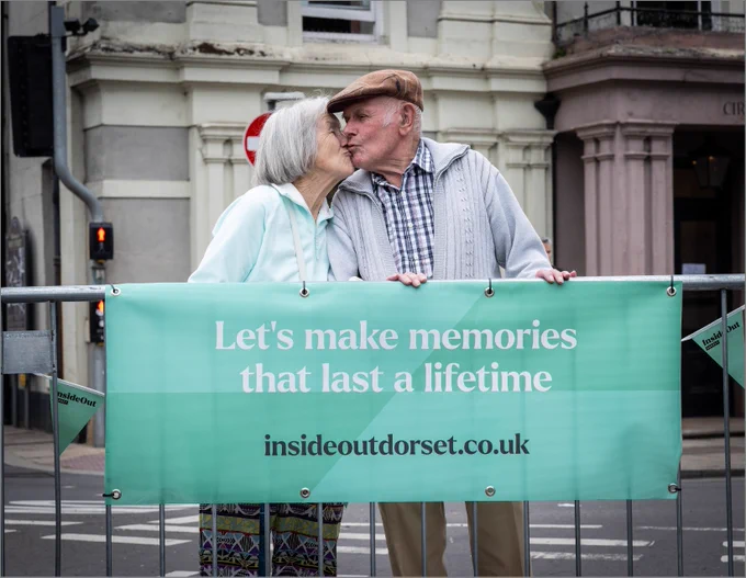 An elderly man and woman share a kiss above a mint green sign that reads: 'Let's make memories that last a lifetime insideoutdorset.co.uk'