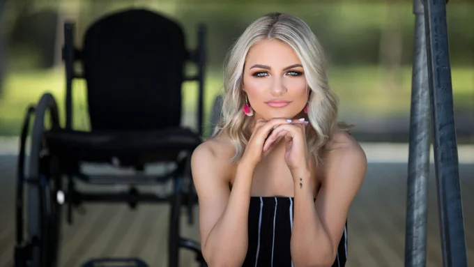 A young blonde woman looks directly at the camera. Behind her, you can see a black wheelchair. On her left wrist is a semicolon tattoo, a symbol of affirmation and solidarity with those who have dealt with suicide, depression, addiction, and other mental health issues.
