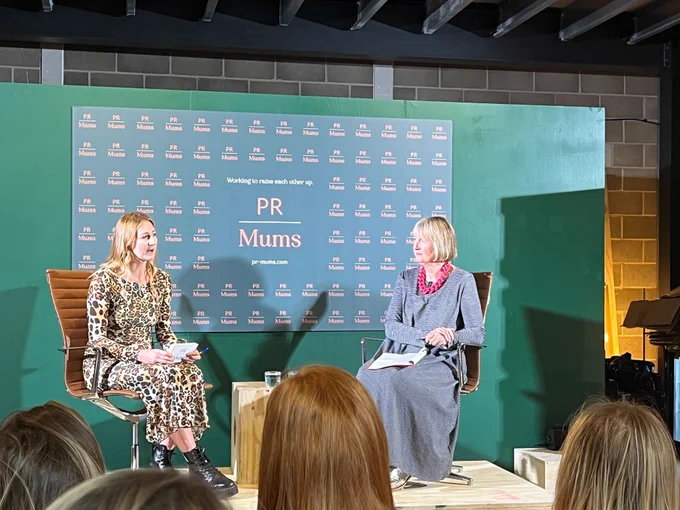 Photo of the speaker stage at the PR Mums breakfast event. Harriet Harman MP sits on the right hand side of the stage in conversation with the moderator. The backdrop shows the PR Mums logo with the tagline "working to raise each other up".