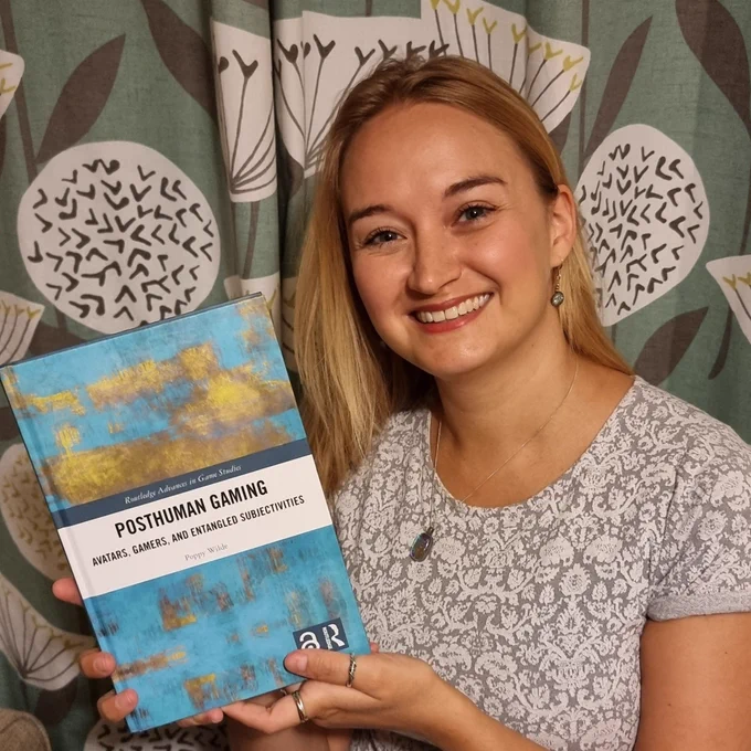 Photo of Dr Poppy Wilde (a White woman in her thirties with long blonde hair, grinning) holding a copy of her book "Posthuman Gaming: Avatars, Gamers, and Entangled Subjectivities".