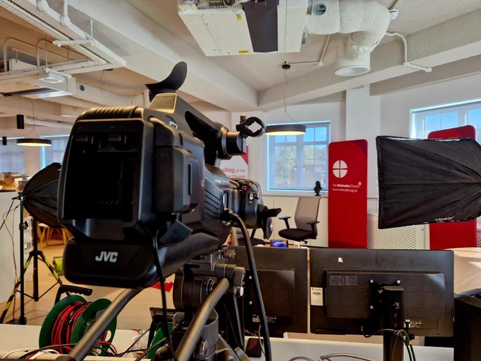 A video camera looking over a desk with monitors, pointing towards a studio area with Methodist branded banners.