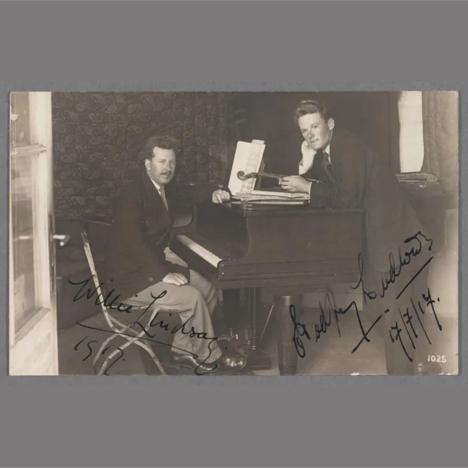 Black and white photographic postcard showing Willie Lindsay sitting at a piano, and Godfrey Ludlow standing with a violin, signed by the respective persons, Ruhleben Internment Camp, Spandau, Germany, 1917. HOLLIS olvwork426123