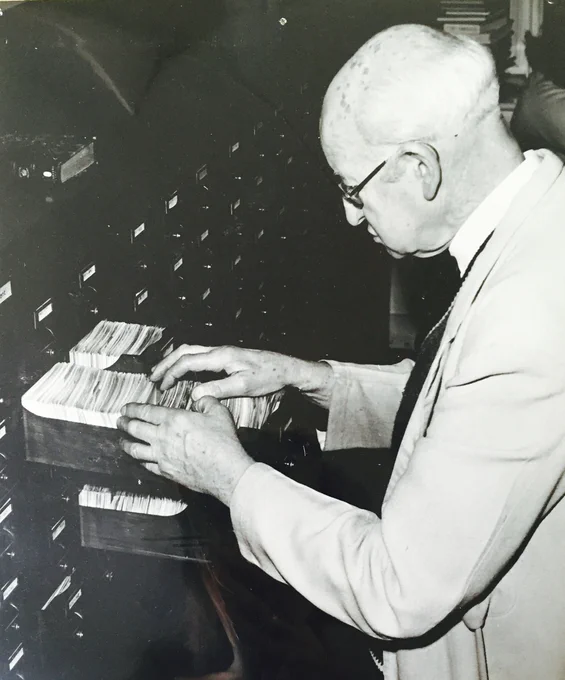 Black and white photo of a man standing in front of a large wooden card cabinet. He has three drawers open and is searching through the cards in one.
