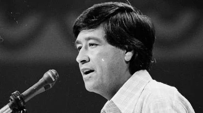 A black and white photo of Cesar Chavez. He is standing in front of a microphone speaking.