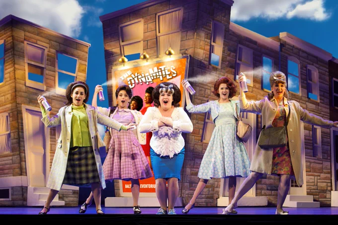 Five actors on a stage with buildings in the background behind them. Four of the actors are holding hairspray cans and spraying them while singing and the actor in the center is singing. They are all dressed in different colored clothing. 