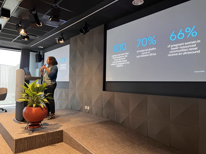 Google Researcher Angelica Willis holding a microphone and speaking at Research@ Accra. Behind Angelica, a presentation slide displays statistics about maternal health.