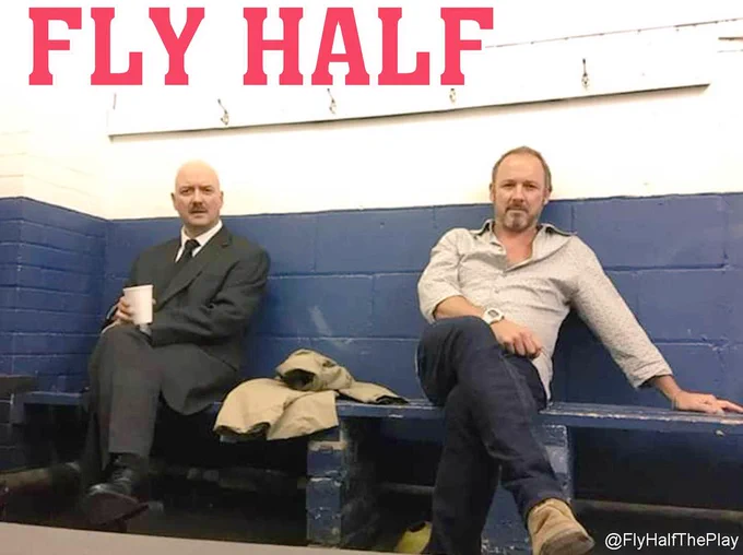 Fly Half writer/actor Gary Lagden and composer/musician Gareth Moulton sit on the bench against a blue wall in a Rugby Club dressing room ready to perform Fly Half The Play - words which appear in red above their heads 