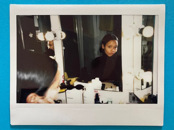 A photo of a polaroid on a blue surface. The polaroid features a young woman in a black jumper looking at the camera via her reflection in a dressing room mirror.