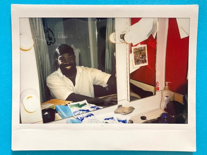 A photo of a polaroid on a blue surface. The polaroid features a young man in a white shirt and black durag smiling at the camera via his reflection in a dressing room mirror.