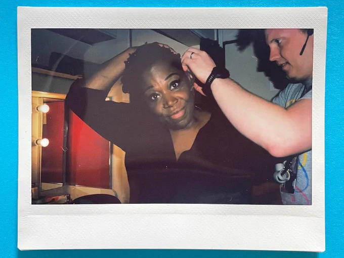 A photo of a polaroid on a blue surface. The polaroid features a woman in a black shirt. She has a small microphone fitted by a man in a grey t-shirt and headset. She smiles at the camera. They are in a dressing room.