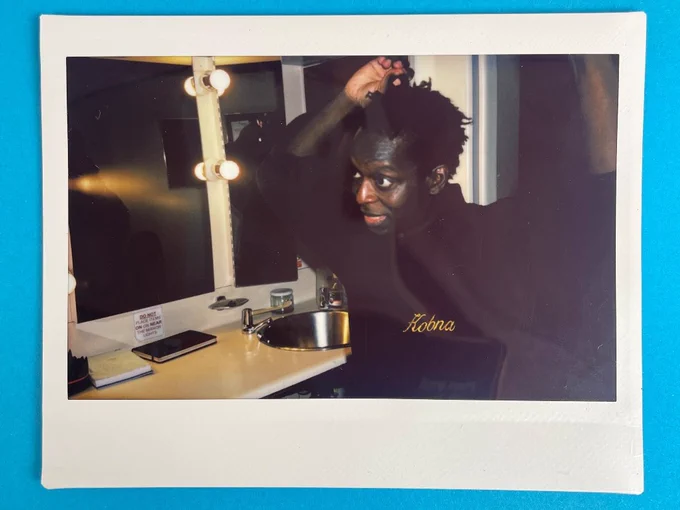 A photo of a polaroid on a blue surface. The polaroid features a man in in a black shirt that has the name 'Kobna' sewn into it in gold. He does his hair whilst looking in the mirror. He is in a dressing room.
