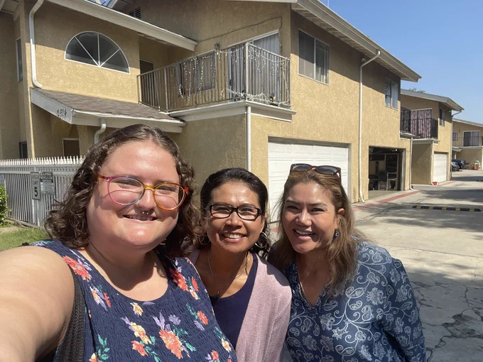 Three people pose for a selfie in front of a block of yellow townhouses. The person on the far left has light skin tone and rainbow glasses. The two women to their left have medium skin tone.