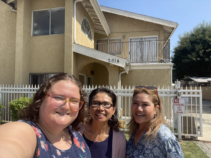 Three people pose for a selfie in front of a block of yellow townhouses. The person on the far left has light skin tone and rainbow glasses. The two women to their left have medium skin tone.