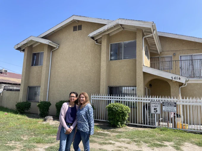 Two women with medium skin tone pose for a photo in front of a yellow townhouse in El Monte. Behind them is a white iron gate with patchy green grass on the floor