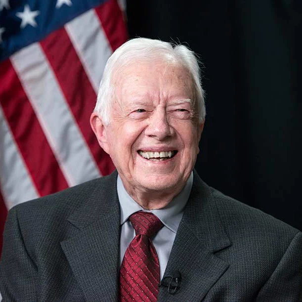 Former President Jimmy Carter smiling to camera in front of an American flag