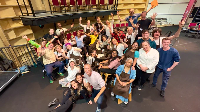 A large group of people gather together on stage and smile and wave at the camera.
