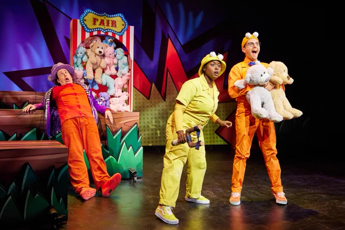 3 actors performing on stage with a large set of stuffed animals behind them. The person on the right of the photo is holding 2 stuffed animals and the person in the center is holding a slingshot.