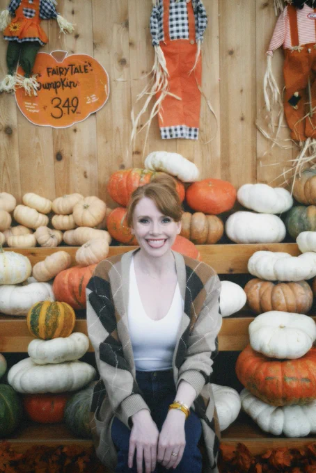 BDH playfully poses in front of a display at Trader Joe’s with pumpkins of all different shapes, sizes, and colors. In full autumn vibes, she wears navy blue jeans, a white t-shirt, and a beige and brown cardigan. 3 smiling scarecrows hang on the wall in the back.