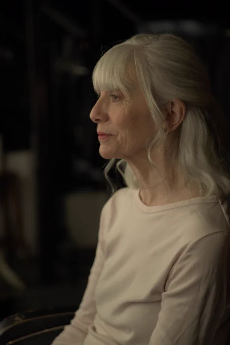 A woman in her sixties stares at herself in a mirror. She is wearing a white long-sleeved top. Her shoulder length hair has loose curls. 