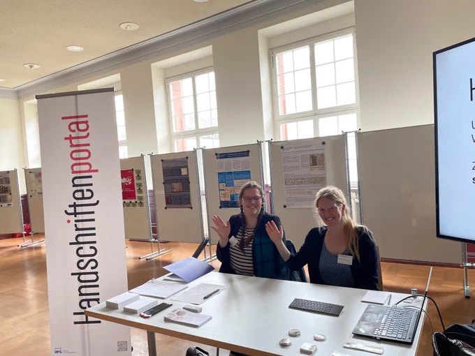 Stand des Handschriftenportals. Zwei Menschen winken in die Kamera.