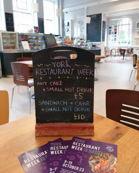 A sign in the York Explore Cafe, saying "York Restaurant Week. Any cake and small hot drink £5. Sandwich and cake and small hot drink £10." with leaflets on the table in front of it.