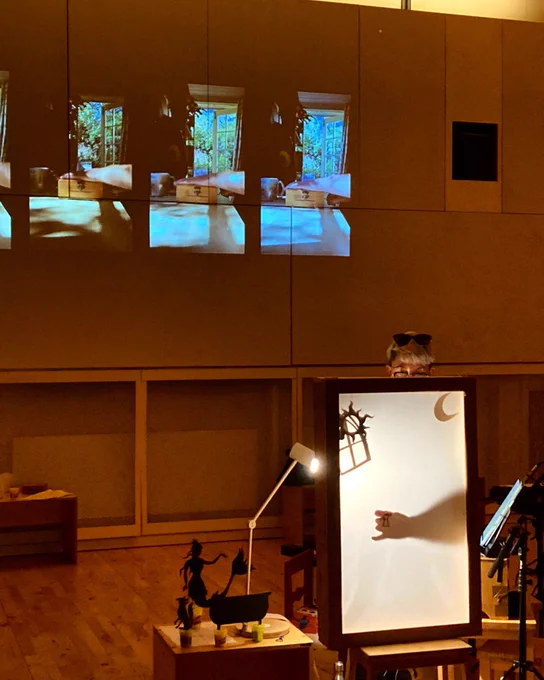 A shadow puppetry lightbox with figures of a moon, sun, and window. Behind, projections of a music box being cranked.