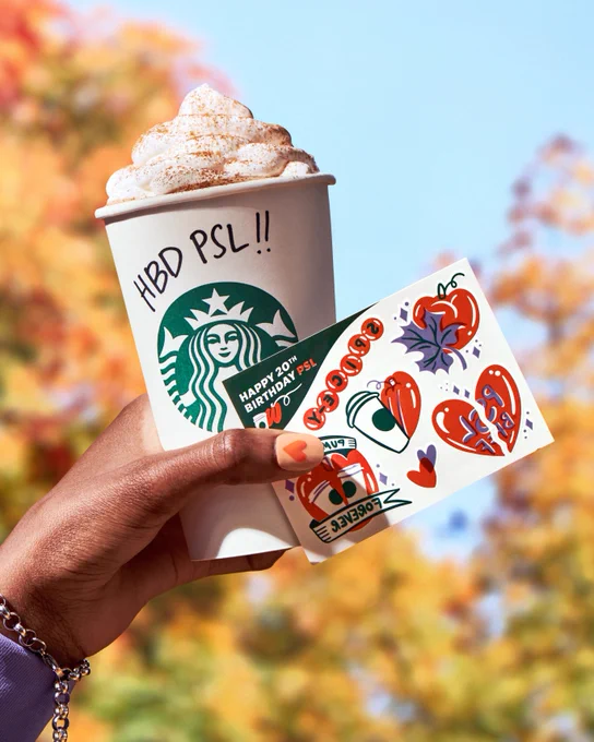 A person holds up a set of custom Starbucks temporary tattoos and a Pumpkin Spice Latte with “HBD PSL!!” written on it against fall foliage.