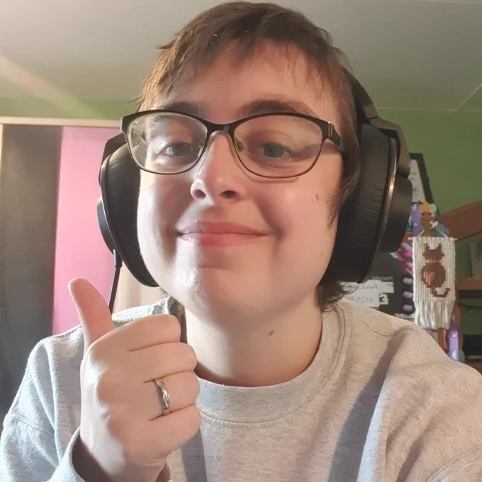 a selfie of Artie smiling at the camera with their thumb up. Artie is a white person with short brown messy hair, wearing glasses, black headphones, and a light grey jumper