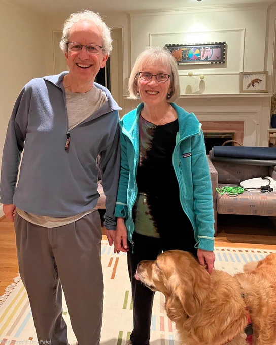 Lawrence Katz (left) and Claudia Goldin (right) standing in their living room with their golden retriever standing next to them.