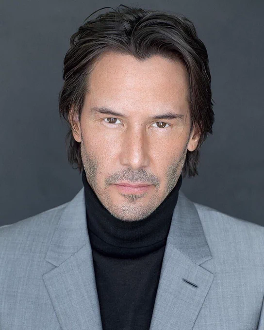 keanu, grey suit, black turtle neck, black hair parted down the middle, going just past his ears, grey five o clock shadow. grey background. 