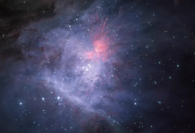 An image of a young star-forming region filled with wispy blue, grey, green, and red nebulosity that is brightest towards the centre and fainter towards the edges, especially in the top left corner and on the right side. Thousands of stars are seen sprinkled across the field, concentrated towards the centre, and the brightest stars show the eight spikes due to diffraction that are characteristic of Webb images.