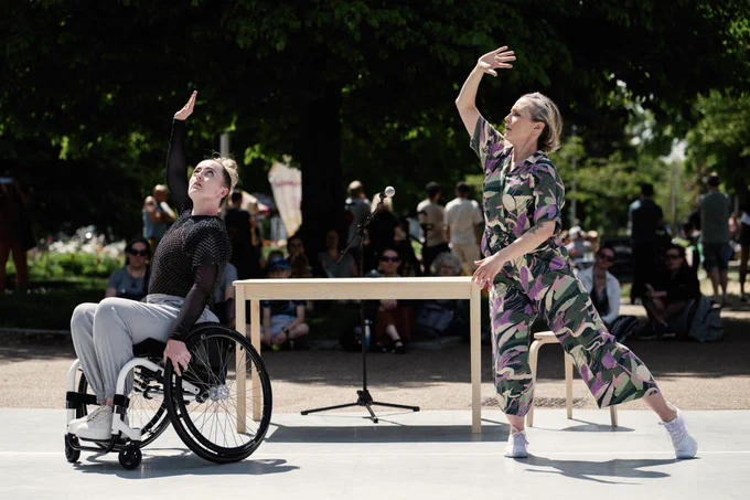 Maiya Leeke and Caroline Lofthouse performing ‘I think we should start over’ at Brighton Festival. They have their left arm lifted above their head and are dancing outdoors on a large grey mat. Behind them there is a rectangular wooden table and microphone stand and the audience is in the background watching the performance.