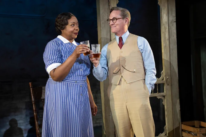 Two actors standing on the set of a play. They are each holding a glass of tea and they are wearing unique outfits.