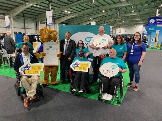 The Euan's Guide team and the Motability Operations CEO and mascot smile to camera with Euan's Guide props at the Big Event in Edinburgh. Some people stand and some people sit in wheelchairs. 