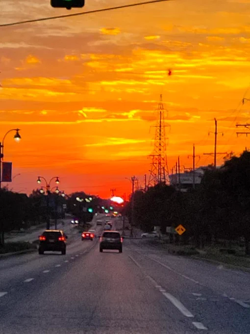 The sum setting ahead of the road with cars driving towards it and street lights on one side and electrical poles on the other.