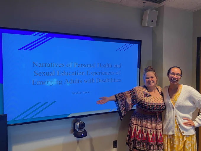 Maddie and Rose smile in front of a screen with Maddie's thesis presentation, titled "Narratives of Personal Health and Sexual Education Experiences of Emerging Adults with Disabilities"