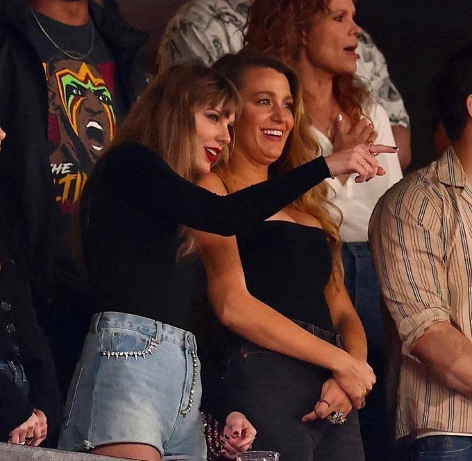 Taylor Swift leans in to talk to Blake Lively at an event. Taylor points to something off camera as Blake smiles attentively.