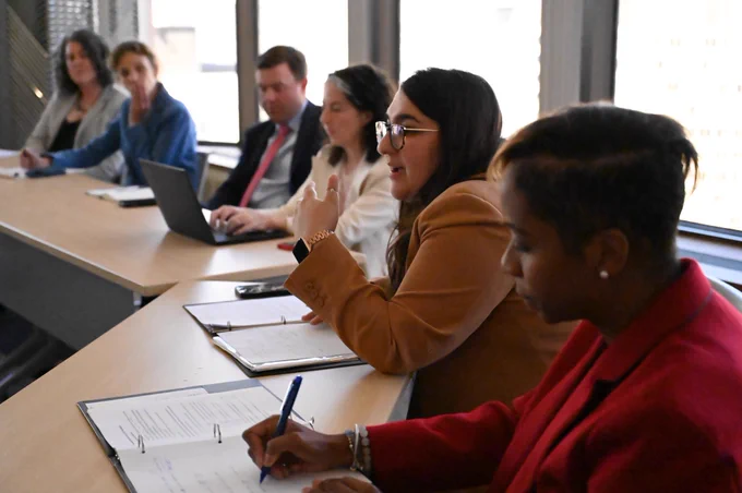 Sapna sitting at a table next to AG Campbell and speaking at a round table.
