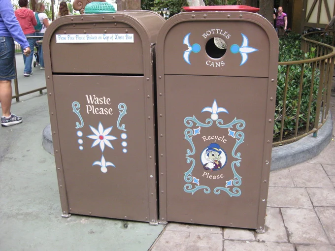 Brown trash cans from Disneyland's Fantasyland with a temporary sign attached that says "Please place plastic baskets on top of waste bins"