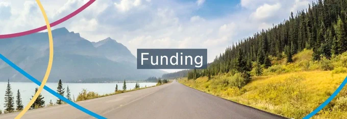A paved road lines a body of water and a strip of coniferous trees. Across the body of water, there are mountains. There are clouds in the blue sky. In the centre, there is a text box containing the word Funding.