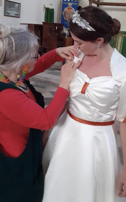 a white blonde costume supervisor fits a white dress with orange trim onto an opera singer who is white with brown hair.