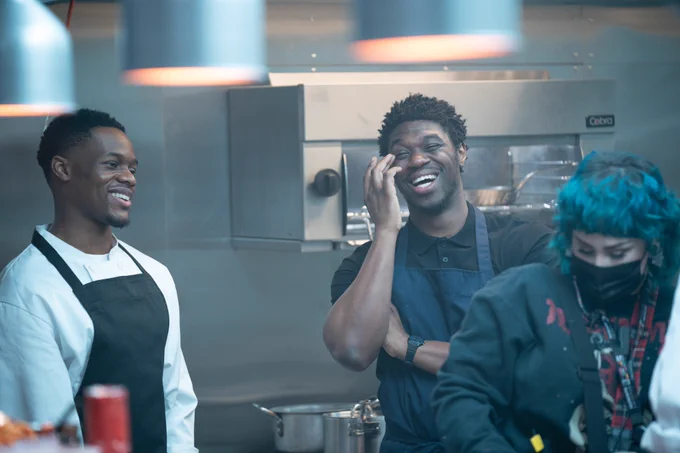 Image shows two actors laughing, they stand in a kitchen and a member of crew is in front of them.