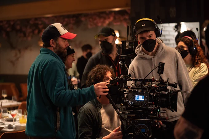 Image shows a director looking at footage on a camera, surrounded by other crew. They are set up in a dark lit restaurant interior.