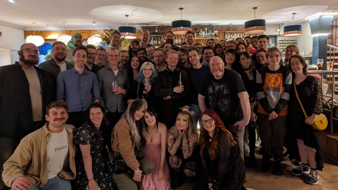 A large group of Yogscast creators, staff, and friends gather for a photo in a warmly-lit bar. They're all smiling for the photo.