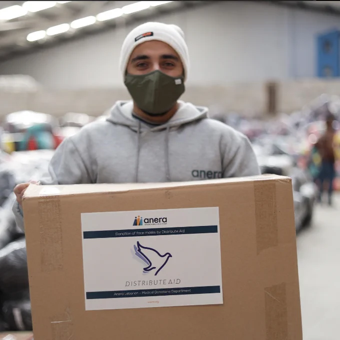 The picture displays a volunteer of our partner organisations holding a box while looking at the camera. the box has the DA logo on it. 