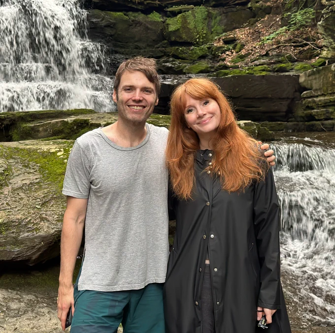 Standing in front of a rushing waterfall and mossy rocks in rural Pennsylvania, Seth Gabel (left) and BDH (right) wrap their arms around each other for a photo. Seth wears a grey t-shirt and teal swim trunks and BDH wears a black long raincoat and her hair down.