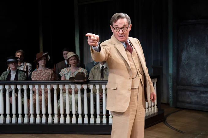 An actor on a stage with several actors in the background. The actor at the front of the photo is pointing in the leftward direction.