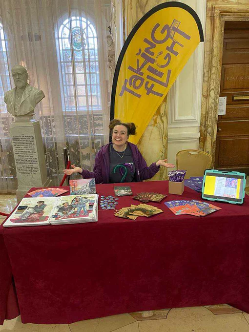 Steph sits behind a table of promotional materials in front of a bright yellow Taking Flight flag in a grand room
