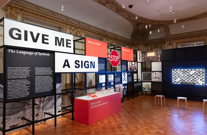 A large white panel says give me a sign in black text and capital letters. There is a small black panel to the left that says "The Language of Symbols" in white text. There are two red panels that ask, "what is a symbol" and "why use symbols." In between the two red panels is a large red stop sign. There are three stools in front of a glowing tv screen with hundreds of black and white symbols. 